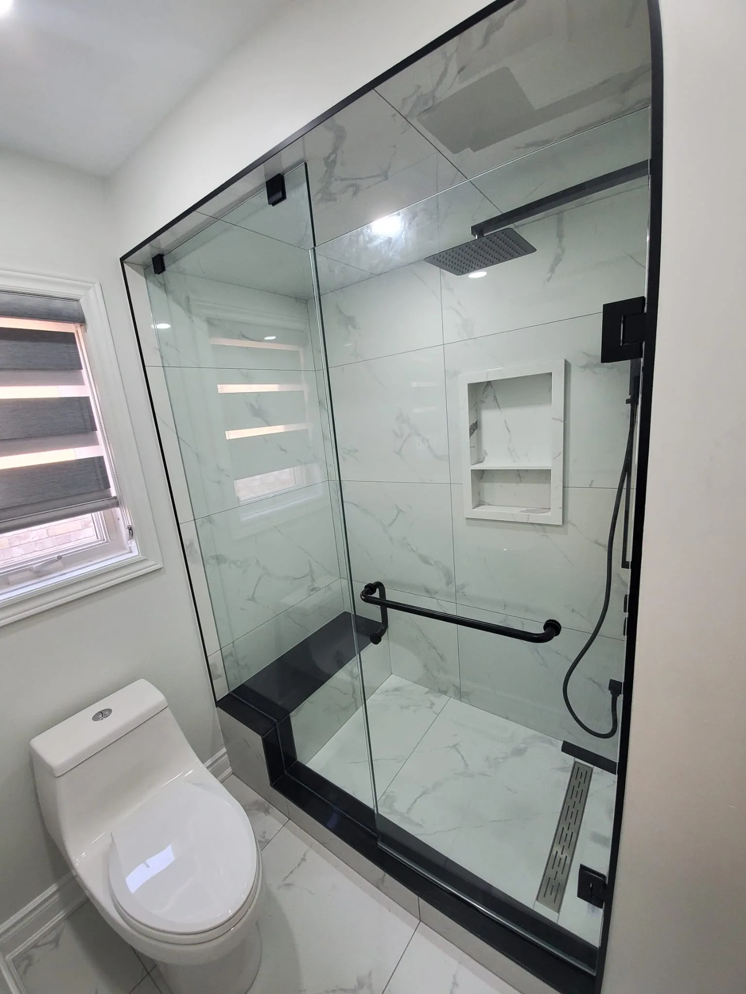Walk-in shower with white marble walls, frameless glass, black bench, and matte black rain head