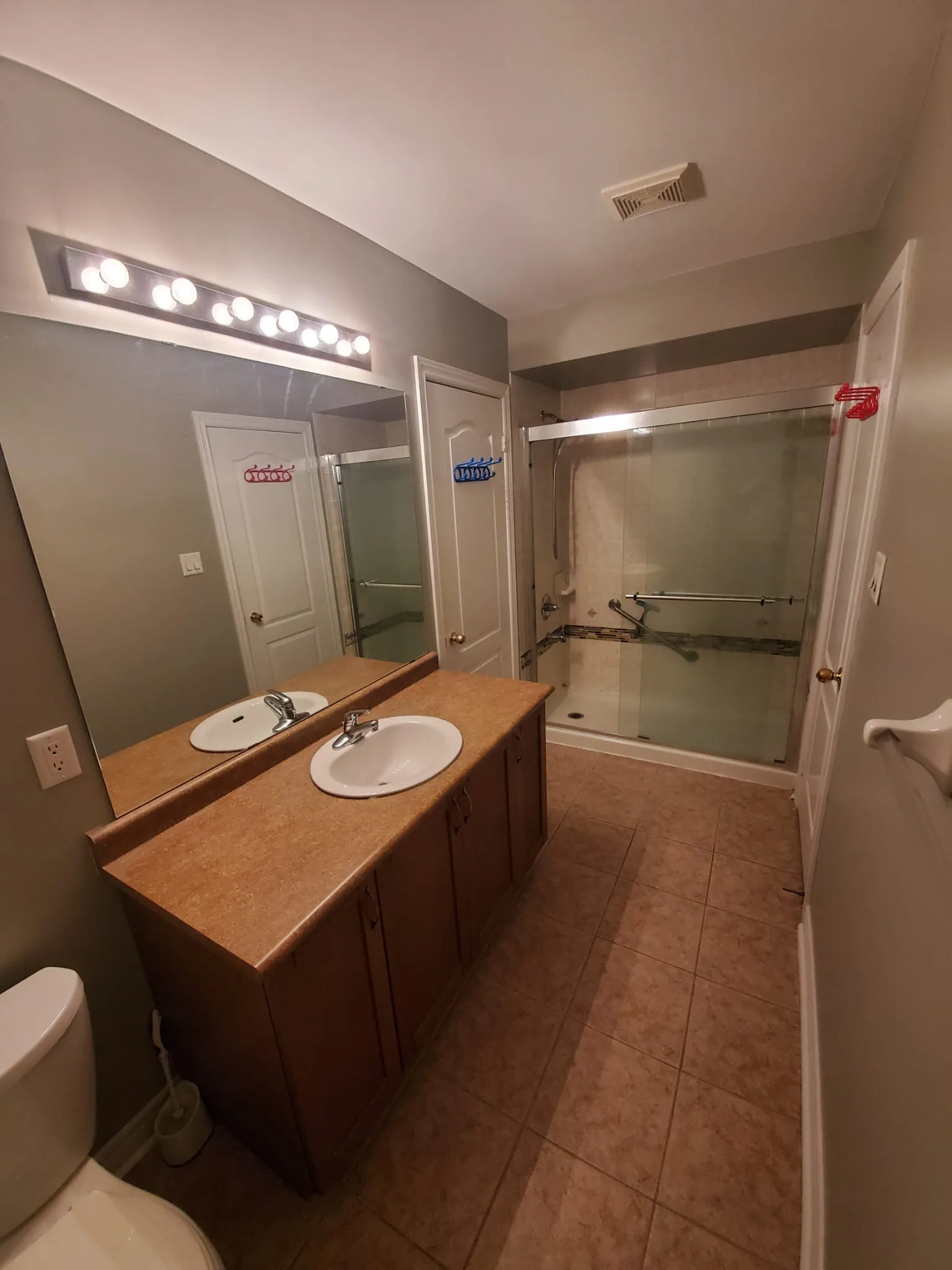 Outdated bathroom with brown laminate vanity, double oval sinks, Hollywood vanity bulbs, and beige tile floor
